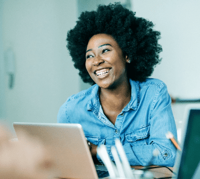 Photograph of a young Black woman with a large afro hairstyle, wearing a denim shirt, smiling broadly with braces visible on her teeth. She is looking happily to her left while seated at a desk with a laptop in front of her.