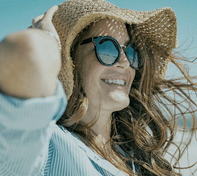 Photograph of a smiling woman wearing a straw hat and sunglasses, looking to the right. Her long, brown hair is blowing in the wind, and sunlight casts dappled shadows through her hat onto her face. She is wearing a light blue striped shirt.