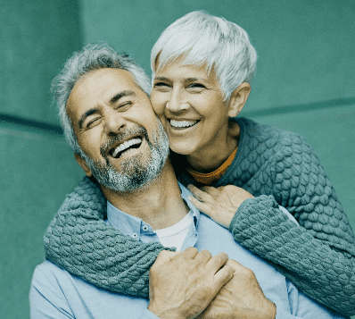 Photograph of a happy older couple. A man with a grey beard is laughing with his eyes closed, while a woman with short white hair hugs him from behind, smiling broadly.