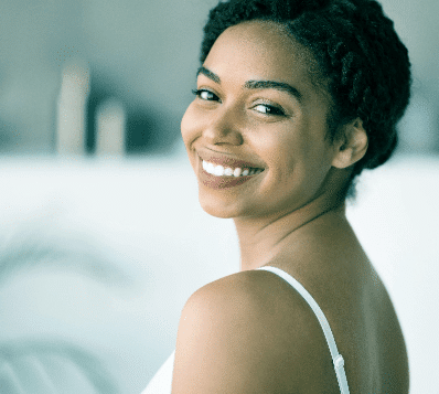 Photograph of a young Black woman with dark, curly hair pulled back, smiling broadly and looking over her left shoulder at the viewer. She is wearing a white strap on her shoulder. The background is blurred and light-colored.
