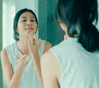 Photograph of an Asian woman applying cream to her face while looking into a mirror. She has dots of cream on her forehead and cheek, and her right hand is spreading it on her chin. Her reflection shows her back and dark ponytail.