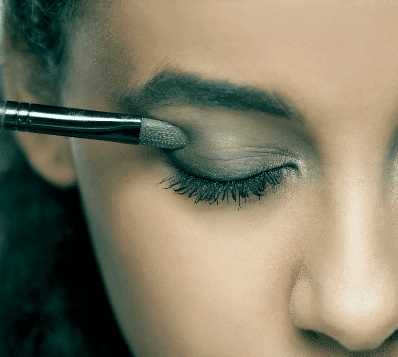 Photograph of a person's closed eye, with a makeup brush applying dark eyeshadow to the eyelid.