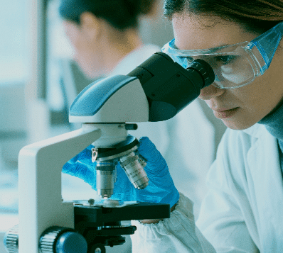 Photograph of a person in a lab coat, blue gloves, and safety goggles looking into a microscope, with another person blurred in the background.