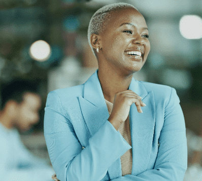 A Black woman with short, light-colored hair, wearing a light blue blazer, smiling broadly and looking to her right. Her left hand is near her chin. A blurred office background is visible.