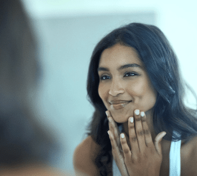 Photograph of a smiling woman with long dark hair looking at her reflection in a mirror while gently touching her chin and jawline with both hands.