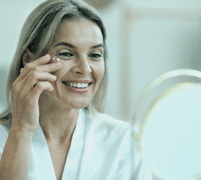 Photograph of a smiling woman with blonde and grey hair, wearing a white robe, applying a small dab of cream under her right eye with her finger while looking into a mirror.