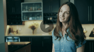A woman with long dark hair and a denim shirt, wearing a lavalier microphone, smiling and looking to the side in a modern kitchen with dark cabinets and under-cabinet.