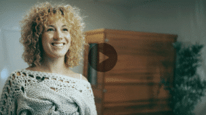 A smiling woman with light brown curly hair, wearing an off-white knitted top. A wooden cabinet and a green plant are in the blurred background.