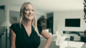 A smiling blonde woman with long hair wearing a black top. She is standing in a modern living room that has white furniture and a wall-mounted TV.