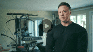 A photo shows a muscular man in a black shirt with a lavalier microphone, standing in a home gym with exercise equipment behind him.